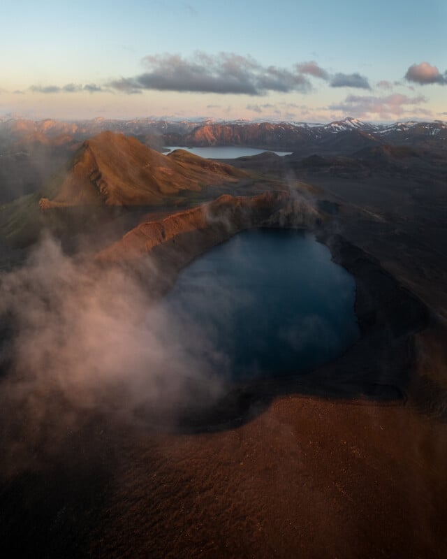 Aerial view of a volcanic crater lake surrounded by rugged, brown mountains with patches of snow and clouds drifting above at sunrise or sunset.