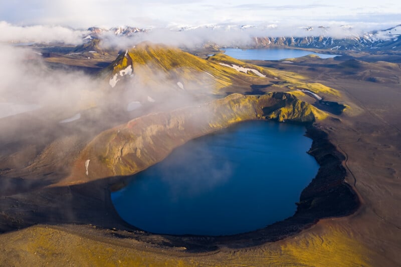 Aerial view of a volcanic crater lake surrounded by rugged, mossy hills and patches of snow, with mist and distant mountains in the background. The landscape appears remote and untouched.