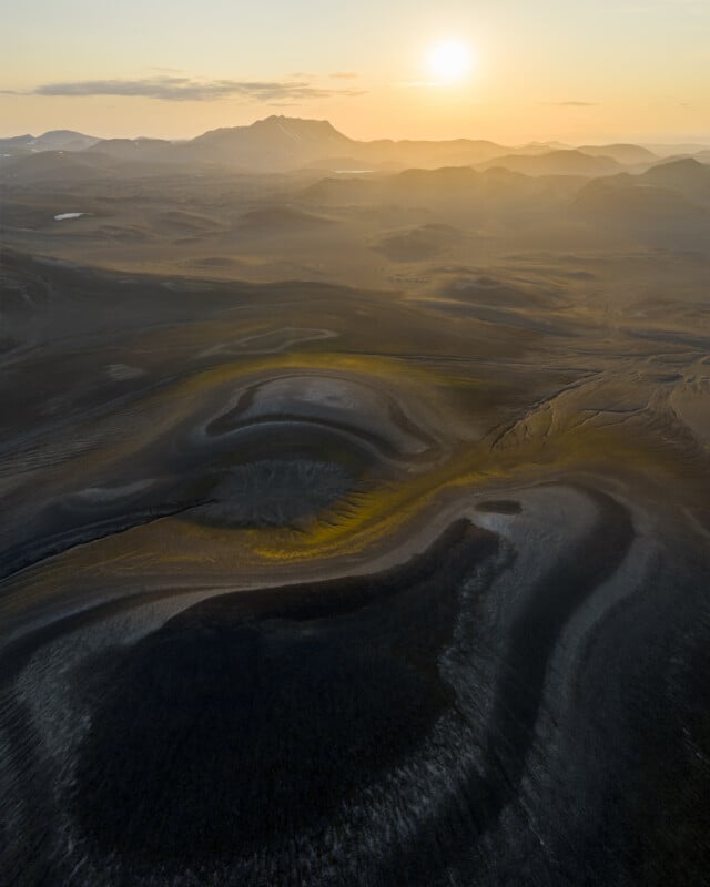Aerial view of rugged, volcanic terrain at sunset, with rolling hills, swirling patterns on the ground, and a distant mountain silhouetted against the glowing sun. Warm light highlights patches of the landscape.