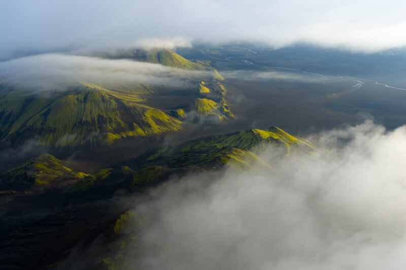 Aerial view of moss-covered mountains partially shrouded in mist and clouds, with a river winding through a dark volcanic landscape in the background.