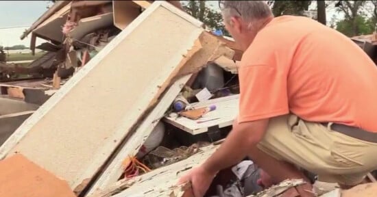 A man in an orange shirt and khaki shorts searches through debris and rubble from a destroyed building, looking for belongings or survivors after a disaster.