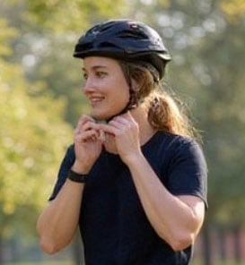 A person wearing a black helmet and a dark shirt is outdoors, smiling and fastening the helmet strap. Trees and greenery are visible in the blurred background.