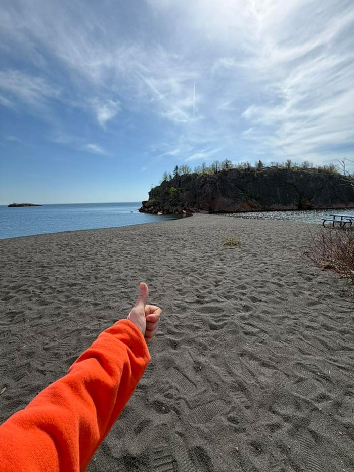 A person wearing an orange sleeve gives a thumbs-up on a sandy beach, facing a rocky outcrop by calm blue water under a partly cloudy sky. A picnic table is visible to the right.