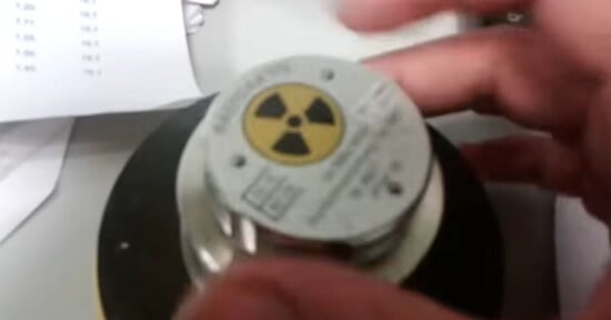 A person’s hand holds a circular metallic object with a yellow and black radiation warning symbol on top, indicating it contains radioactive material. A piece of paper is visible in the background.