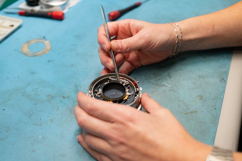 A person uses a metal tool to repair or adjust the internal parts of a camera lens on a blue work surface. Various small tools and camera components are visible nearby.