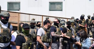 A group of armed police officers wearing tactical gear and face coverings stand in front of a building, while a woman in the foreground holds a camera and interacts with one of the officers.