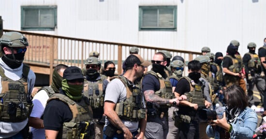 A group of armed police officers wearing tactical gear and face coverings stand in front of a building, while a woman in the foreground holds a camera and interacts with one of the officers.