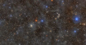 A dense starfield in deep space, featuring countless stars of varying brightness and color, including prominent blue and orange stars, set against a dark background with faint nebula clouds.