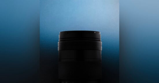 A camera lens in shadow is centered against a gradient blue background, with only the top and textured grip clearly visible.