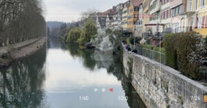 A calm river runs alongside colorful buildings and a stone walkway, where people are seated. Leafless trees line the opposite bank. A camera viewfinder overlay shows settings at the bottom of the image.