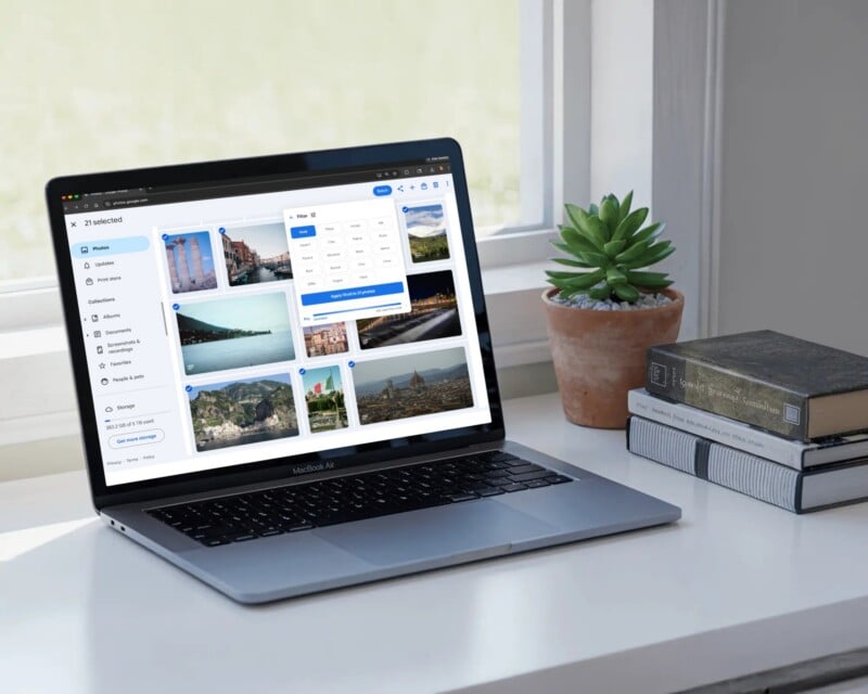 A laptop on a white desk displays a photo gallery and map interface. Nearby are a potted succulent, two stacked books, and a phone, with daylight coming through a window in the background.