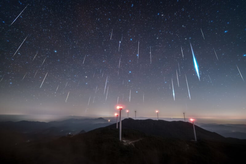 A starry night sky with numerous meteor streaks above distant mountains; several wind turbines with red lights stand on the ridgeline below.