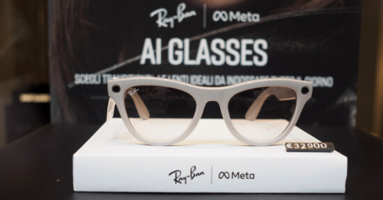 A pair of Ray-Ban and Meta AI glasses is displayed on a white stand with the price tag €329.00. The background features branding for Ray-Ban and Meta with the text "AI GLASSES.