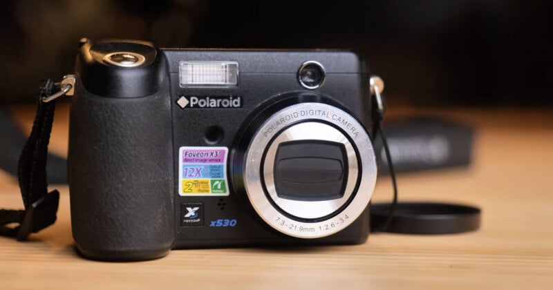A close-up of a Polaroid x530 digital camera with various colorful stickers on the front, placed on a wooden surface with its lens cap off and lying nearby.