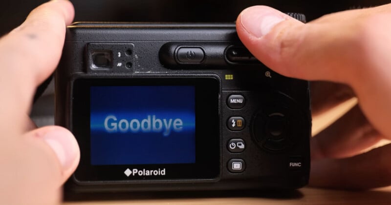 A close-up of a person holding a Polaroid digital camera displaying the word "Goodbye" on its screen, with fingers visible pressing buttons on the camera.