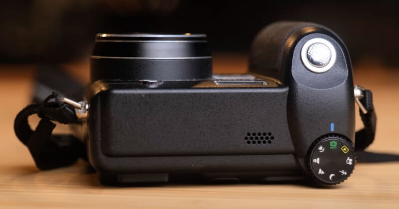 A close-up of a black digital camera laid on its back, showing the mode dial with various settings, the shutter button, and part of the attached lens, with a strap visible on the side.