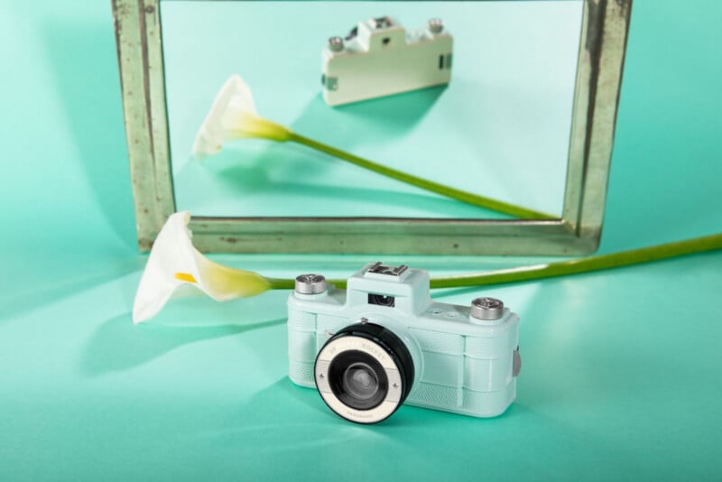 Una cámara vintage azul claro y una cala blanca se colocan frente a un espejo que refleja los dos objetos, todo sobre un fondo turquesa claro.