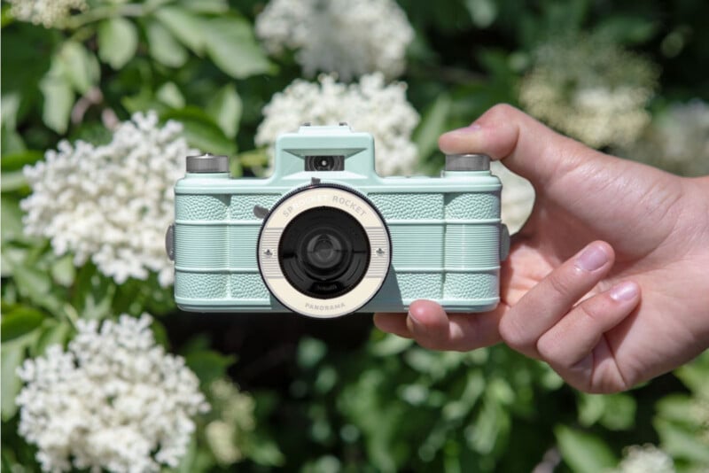 Una mano sostiene una cámara de película vintage de color verde menta pastel al aire libre con flores blancas borrosas y hojas verdes en el fondo.