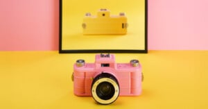 A pink vintage camera sits on a yellow surface with a pink background. Behind it, a mirror reflects the camera, but the reflection appears as a yellow camera instead of pink.