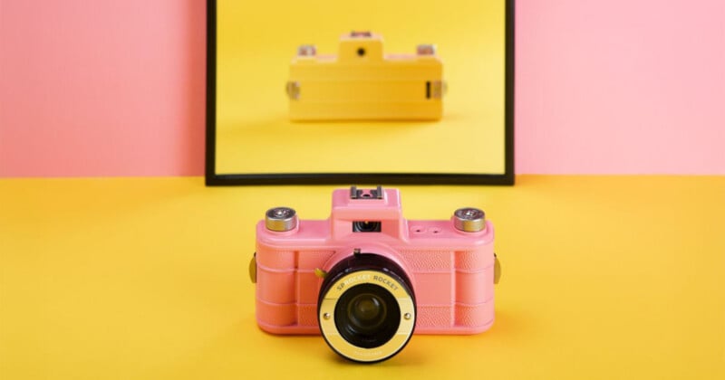 Una cámara vintage rosa se asienta sobre una superficie amarilla sobre un fondo rosa. Detrás, un espejo refleja la cámara, pero el reflejo muestra una cámara amarilla en lugar de rosa.