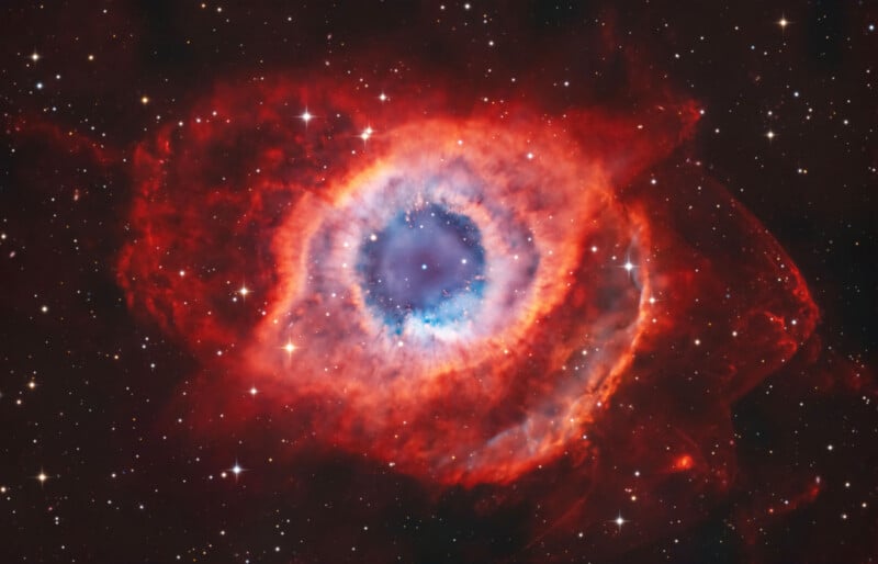 A vibrant image of the Helix Nebula, showing a bright blue core surrounded by vivid red and orange gas clouds, with numerous stars scattered in the dark background of space.