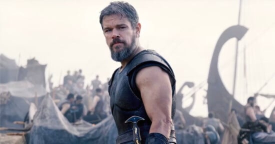 A bearded man in medieval armor stands in front of a busy camp with tents and people, holding a sword at his side, with ships in the background.