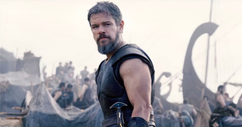 A bearded man in medieval armor stands in front of a busy camp with tents and people, holding a sword at his side, with ships in the background.