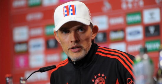 A man in a white cap and black and red sports jacket with a Bayern Munich logo speaks at a press conference, with a red background featuring various sponsor logos.