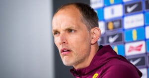 A man with short hair and a trimmed beard, wearing a maroon jacket, looks thoughtfully ahead during a press conference. A blurred background shows various sponsor logos on a wall.