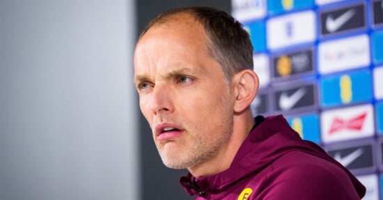 A man with short hair and a trimmed beard, wearing a maroon jacket, looks thoughtfully ahead during a press conference. A blurred background shows various sponsor logos on a wall.
