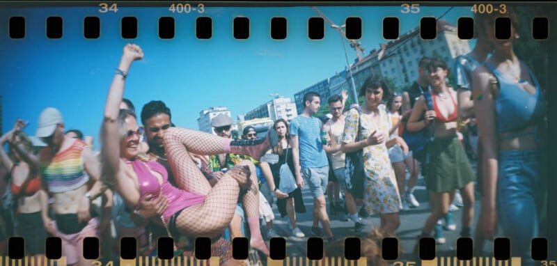 Un grupo de personas vestidas con trajes coloridos desfilaron al aire libre. Un hombre vestido con ropa rosa y medias de red es sostenido por otro hombre y sonríe. Bajo el cielo azul, me siento feliz y feliz.