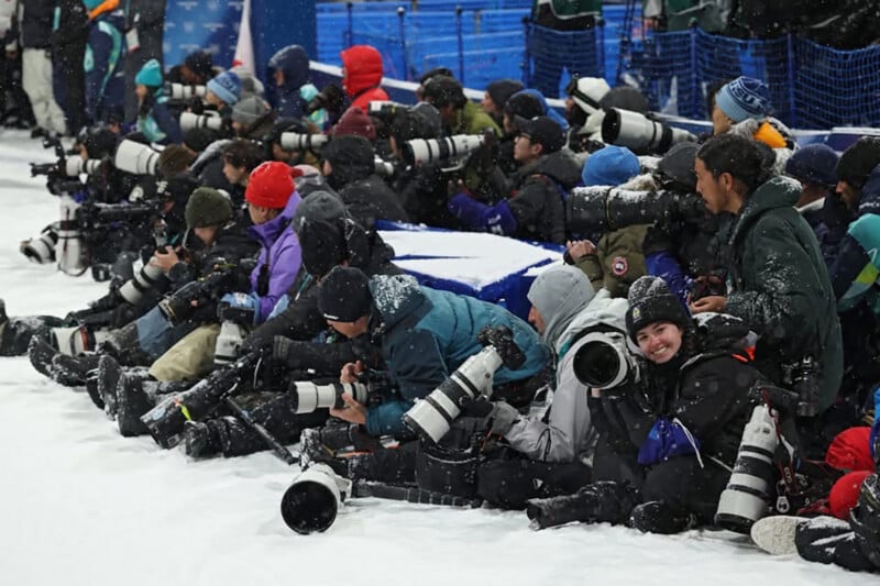A group of photographers bundled in winter gear crouch in the snow, aiming large telephoto cameras, likely at a sporting event. Snow covers the ground and some camera equipment.