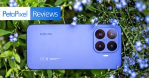 A blue Xiaomi smartphone with triple camera lenses lies among green leaves and small blue flowers. The image has a "PetaPixel Reviews" label in the top left corner.
