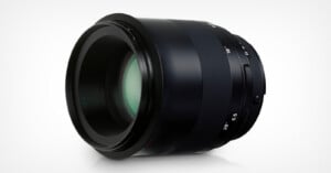 A black camera lens with a wide glass front element, visible focus markings, and a smooth, metallic finish, photographed against a plain white background.