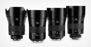 Four black Zeiss Otus camera lenses of different focal lengths (28mm, 55mm, 85mm, and 100mm) are standing upright in a row on a white background.