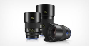 Three Zeiss Otus camera lenses are displayed on a white background; two stand upright with lens hoods attached, and one is lying on its side showing the front element and blue Zeiss logo.