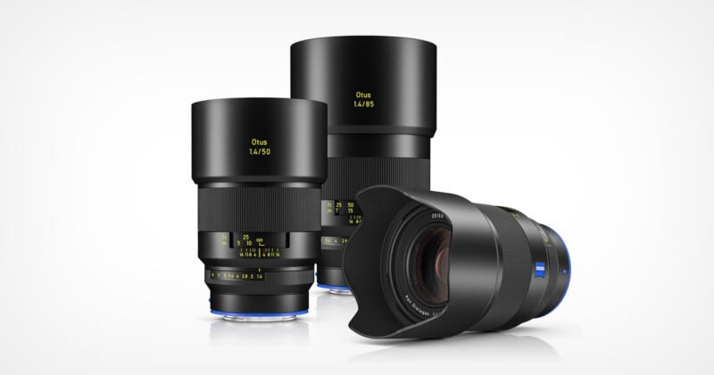 Three Zeiss Otus camera lenses are displayed on a white background; two stand upright with lens hoods attached, and one is lying on its side showing the front element and blue Zeiss logo.