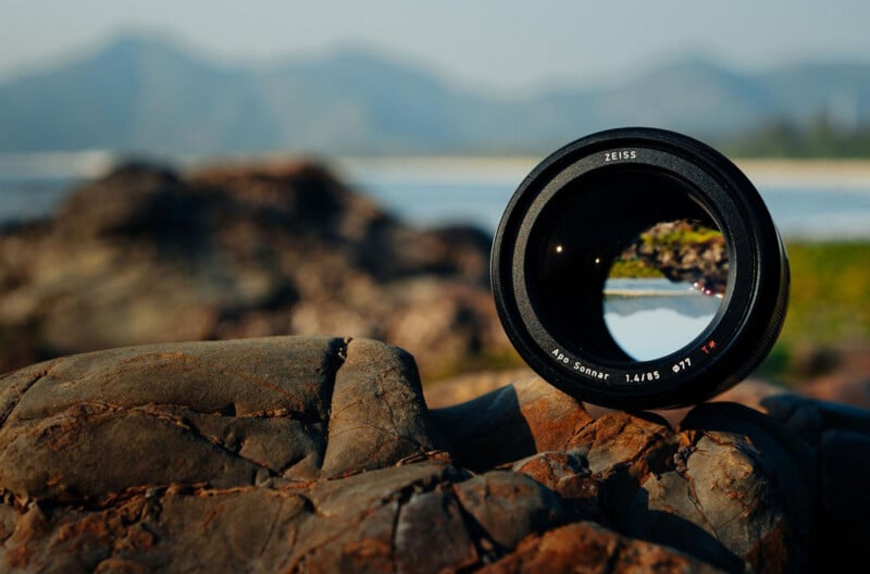 A camera lens rests on rocky ground, with a scenic landscape of mountains and water visible in the background. The view through the lens sharply shows the distant landscape in focus.