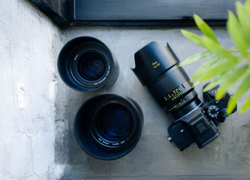 A black DSLR camera with a large lens is placed next to two additional camera lenses on a concrete surface near a window, with a leafy green plant partially visible on the right.
