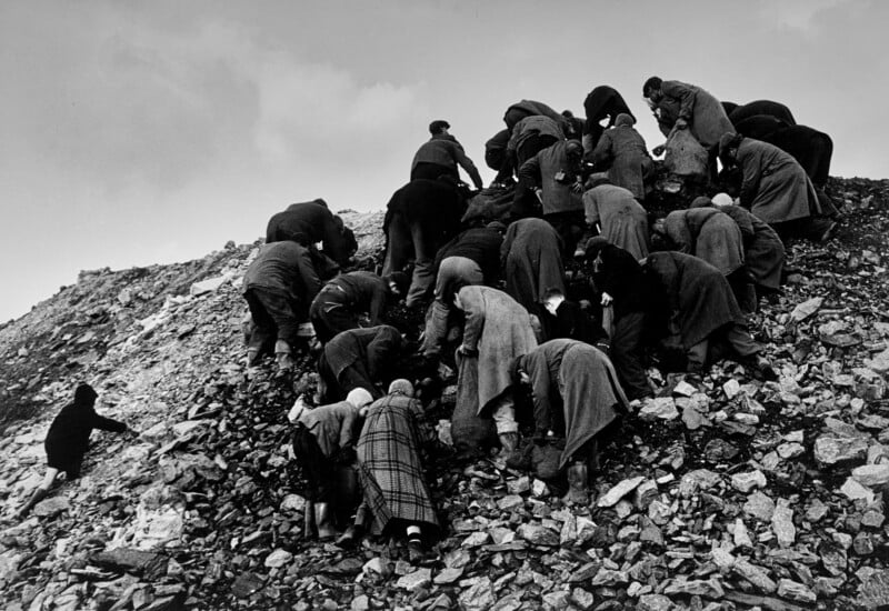 A group of people wearing coats climb and search through a rocky hillside under a cloudy sky, appearing to look for something among the stones.