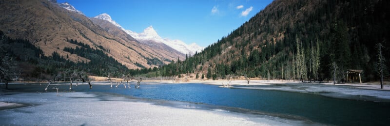 A partially frozen lake sits before a valley of snow-capped mountains and dense pine forests under a clear blue sky.