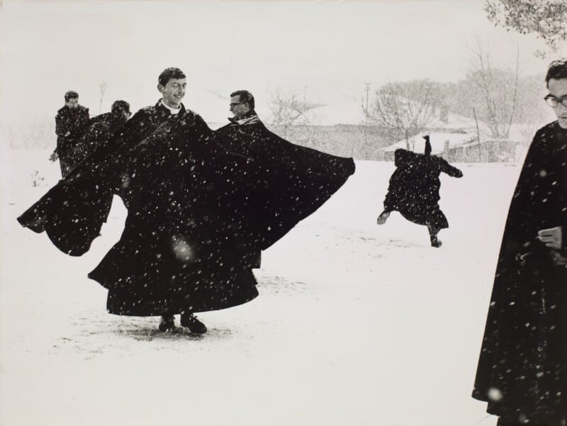 Several men in long black cassocks play in the snow, with one figure leaping in the background and another standing in the foreground.