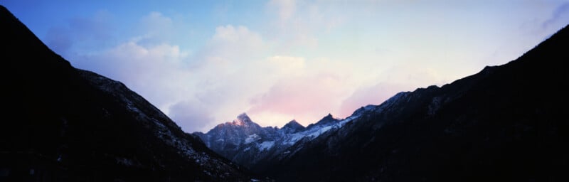 A panoramic view of snow-capped mountain peaks at sunrise, framed by dark, silhouetted slopes under a soft, pastel-colored sky.