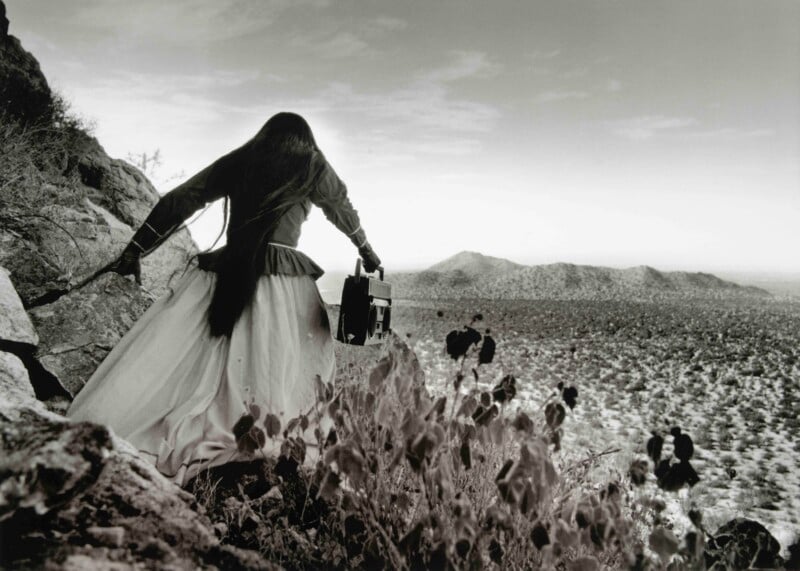 A person with long dark hair, wearing a long skirt and top, stands on a rocky hillside holding a boombox while looking out over a desert landscape.