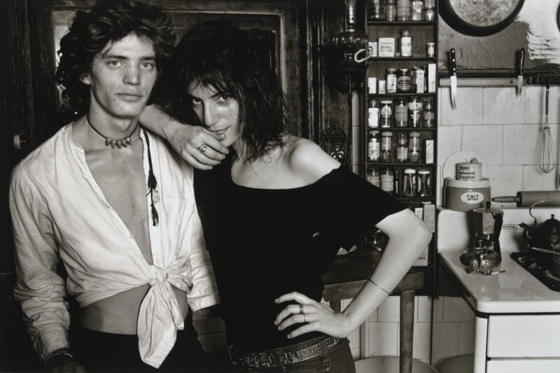 A young man and woman pose together in a kitchen, with the woman leaning her arm on the man's shoulder.