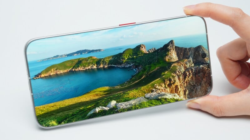 A hand holds a smartphone displaying a landscape photograph of a green, rocky coastline and a blue bay.