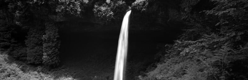 A black and white panoramic view of a waterfall cascading over a dark, recessed cliff face surrounded by dense forest.