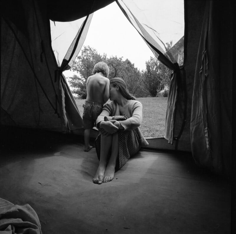A woman sits inside a tent entrance, looking outside, while a shirtless young boy stands beside her, facing away. Grass, trees, and a house are visible in the background.