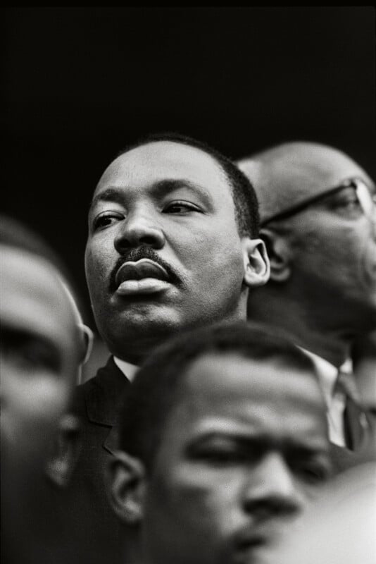 A close-up, black-and-white portrait of Martin Luther King Jr. looking off to the side, surrounded by other men in a crowd.