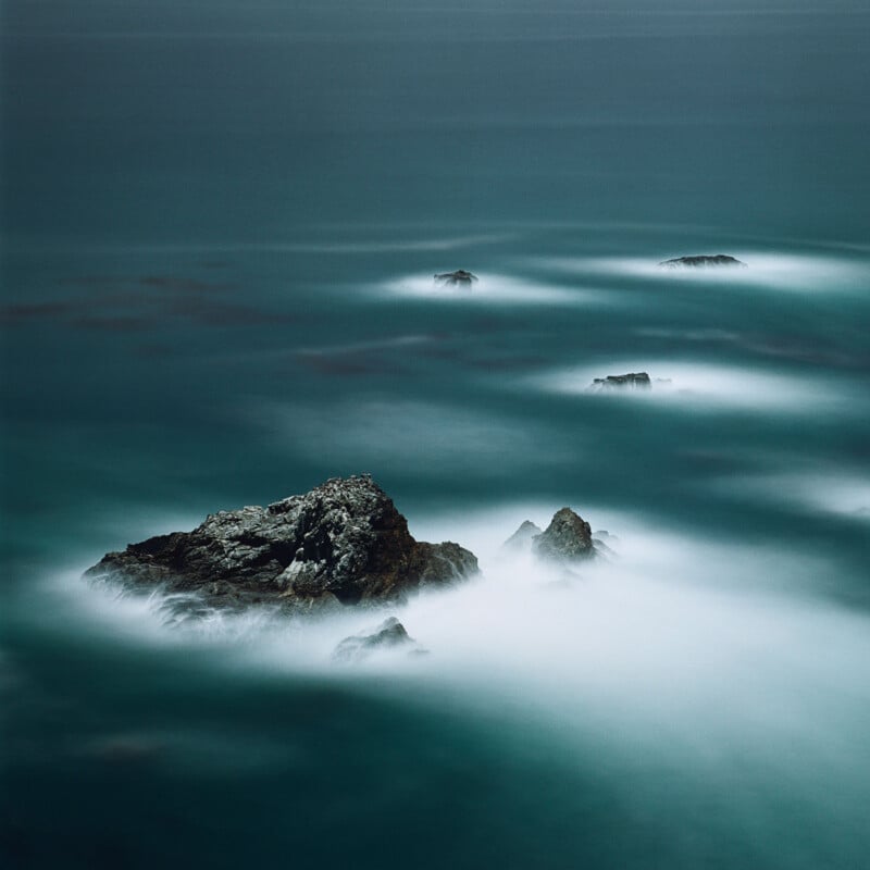 Rocky outcrops rise from a turquoise sea, partially shrouded by mist. The water appears smooth and hazy, creating a dreamlike, tranquil atmosphere under a soft, overcast sky.